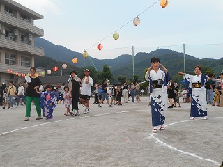 グラウンドで浴衣姿の二人の女性が盆踊りを踊り、その周りには提灯が飾られ、多くの人々が集まってお祭りの雰囲気を楽しんでいる写真