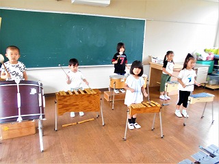 フローリングの室内で、6人の園児たちが大太鼓、木琴、鉄琴などの様々な打楽器を演奏している音楽活動の様子の写真