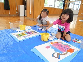 室内で二人の女の子が床に敷かれた青いビニールシートの上で、それぞれカラフルな絵の具と筆を使い、大きな紙に絵を描いている写真