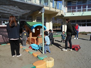 園庭で、黒い服を着た保護者と思われる大人が2人、その周りで数人の子供たちが砂場と遊具のスペースで遊んでいる様子の写真