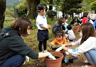 屋外でマスクを着用した複数の大人や子供たちが一緒にしゃがみ込み、植木鉢に小さな苗を植え付けている様子の写真