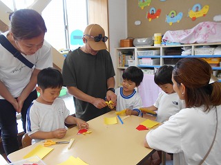 明るい室内で、保護者が見ているなか、体操服姿の園児たちが椅子に座り、黄色や赤色の色紙やハサミが置かれた机の周りで折り紙を楽しんでいる様子の写真