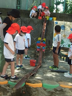 屋外の砂場で赤と白の帽子をかぶった複数の園児たちが、傾斜をつけた樋にバケツから水を流し込んで遊んでる園児や、オレンジと緑の丸い石が交互になっている道を渡ろうとしている園児の写真