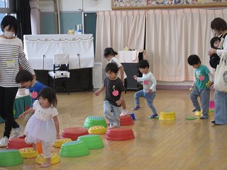 保育園の室内で、園児たちがカラフルな飛び石のような遊具を床に並べた平均台の上を、バランスを取りながら楽しそうに歩いて渡る運動遊びを、数名の大人たちが見守っている写真