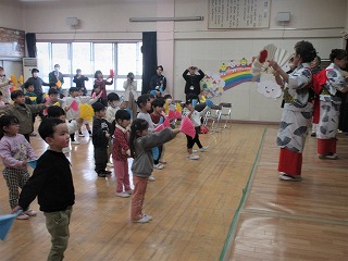 大勢の園児たち広い部屋で、着物姿の大人から両手に持った扇子のような飾りを使って踊りを教わっている様子の写真