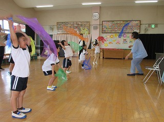 広々とした木の床のプレイルームのような部屋で、体操服のような白いシャツと紺色のハーフパンツを着用した複数の幼児が、紫やオレンジ、青、緑といった様々な色の薄い布を両手に持って頭上に上げたり、下げたりしている写真