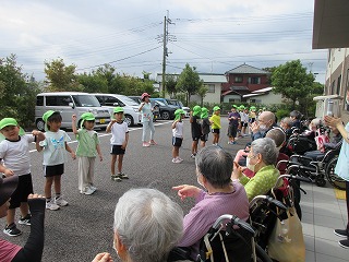 曇り空の屋外駐車場のような場所で、緑色の帽子をかぶった複数の子供たちが、手を上げダンスを披露し、それを見物席のように並んで車椅子に座っているお年寄りたちが、拍手をしたり指をさしたりしながら楽しそうに鑑賞している、交流イベントの様子の写真