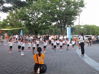 広々とした公園の広場で、体操服を着用し、スカーフを首に巻いた複数の幼稚園児が、周りを多くの観客に囲まれ、指導者の女性の指示に合わせて手を広げて手話を披露している様子の写真