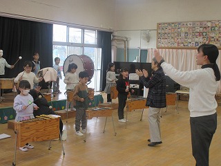 広々とした室内で、園児が木琴やドラムなどの楽器を演奏する合奏の練習をしており、画面右側には両手を上げてリズムを取る白いセーター姿の女性教師と、手を叩く別の女性教師が指導する様子の写真