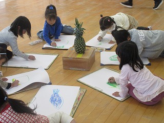木の床の広い部屋で、複数の子供たちが床に広げた白い画用紙とクリップボードに向かい、クレヨンを使って絵を描いており、その中央には木箱の上に置かれたパイナップルをモチーフに写生している様子の写真