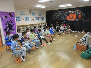 ハロウィーンの飾り付けがされた黒い幕の側で園児たちが小さな椅子に一列に座り、それぞれ紙のようなものを持っており、向かい合わせで正座している女性の先生が話をしている様子の写真