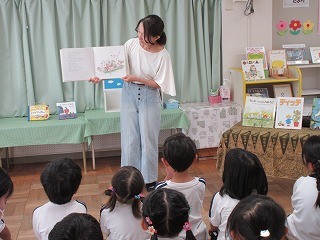 幼稚園の教室で、白いトップスと薄いブルーのジーンズを着用した女性が絵本を広げて、床に座っている複数の子供たちに両手で開いた大きな絵本を掲げながら、読み聞かせをしている様子の写真