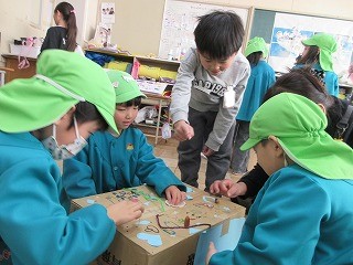 教室で、緑色のスモックと帽子をかぶった子供たちが、段ボール箱の上貼られた木の実や紙片などの素材を使って作ったボードゲームらしきもので遊んでいるのを小学生が説明している写真