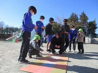 園庭で、青いジャージや黒い服を着た中学生の数人の生徒が、手に持った紐のようなものを操作しながら、地面に置かれたカラフルな板の上にしゃがみ込む園児が駒を回しているのを見守っている写真