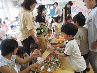 幼稚園の教室で、複数の幼児と保護者、未就園児が賑やかに集まっており、画面手前では白いTシャツ姿の男の子が網の上に手作りの焼鳥を使ってお店屋さんごっこをして未就園児が買いに来て遊んでいる写真