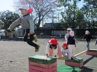 快晴の園庭で、赤と白の帽子をかぶった複数の園児たちが、跳び箱と跳び箱に板を渡らせ橋のようにした所を渡り、3段の跳び箱の上で一人の男の子がダイナミックに飛び跳ねる様子を捉えた写真