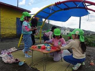 園庭の遊具の下で、緑色の帽子をかぶった複数の園児たちがござの上で小さなテーブルを囲み、おままごとやお店屋さんごっこで遊んでいる様子の写真