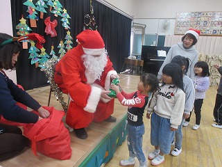 室内のステージ上で、サンタクロースに扮した男性が園児にプレゼントを手渡しており、数人の園児たちが列を作ってプレゼントを貰っている写真