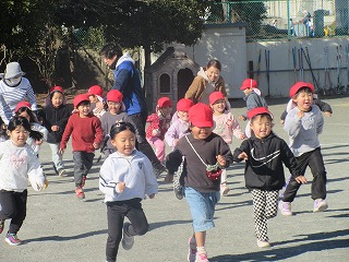 快晴の園庭で赤い帽子をかぶった十数名の園児たちが、数人の先生と一緒に満面の笑顔で元気いっぱいに駆け回る写真