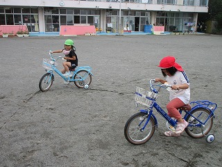 園庭の土の上で、赤と緑の帽子をかぶった2人の園児が、それぞれ青い補助輪付きの自転車に乗って練習している様子の写真