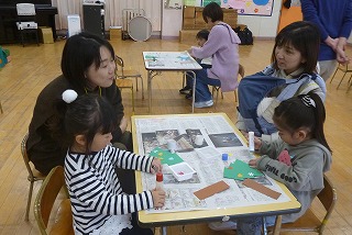 保育園の室内で、親子制作のような活動が行われており、2組の親子がテーブルを囲み、母親たちは楽しそうに話しながら、子どもたちは新聞紙の上に広げた紙工作に集中して取り組んでいる写真