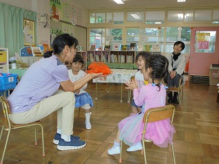 保育園の広々とした室内で、紫のトップスを着た女性教師が、オレンジ色の物体を手に持ち、椅子に座っている2人の女の子の園児と笑顔で会話をしており、その様子を別の女性教師が後ろから見守っている写真