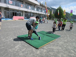 園庭で、体操服姿の男性が、緑の帽子をかぶった園児に、緑のマットの上で相撲のような組み方の指導をしており、周囲を他の園児や先生たちが見守っている写真