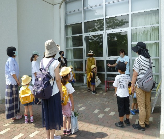 大きなガラス窓のある建物の入口前で、黄色い帽子とリュックを背負った幼稚園児らしき子どもたちと、マスクや帽子を着用した数名の保護者たちが、保育士と思われる女性から説明を受けている様子の写真