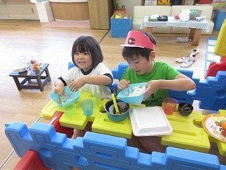保育園の教室内で、大きなブロックで作られたテーブルと椅子に座り、キャップをかぶった男の子と隣の女の子が、お椀に入った泥や草、木の実などを調理しているように見立てて楽しそうにごっこ遊びをしている写真