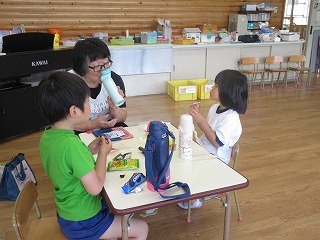 保育園の教室内で、緑のTシャツの男の子と体操服姿の女の子の園児2人が、黒いエプロン姿の女性教師と一緒に、小さなテーブルを囲んで制作活動に取り組んでいる様子を写した写真
