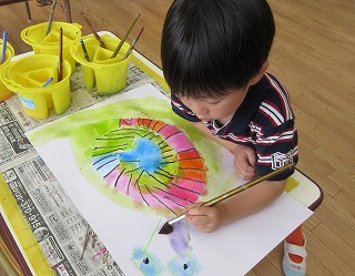 園児の男の子が、新聞紙を敷いたテーブルの上で、水彩絵の具を使い、青、ピンク、オレンジ、緑などの鮮やかな色で、渦巻き模様の抽象画のような絵に集中して筆を走らせている写真