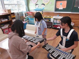 保育園の室内で、胸当て付きの鉄琴を着用した2人の女の子と、その鉄琴を支えている女の子が、一緒に楽器の演奏を楽しんでいる様子の写真