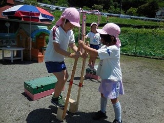 園庭で、ピンク色の帽子をかぶった体操服姿の2人の園児が、木製の足踏み式竹馬を使って遊び、もう一人の園児がその様子を後ろで見守っている写真