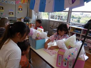 保育園の室内で行われている夏祭りのような行事で、浴衣を着た女の子が、ビニール袋に詰められた綿菓子風の模擬商品を、お客さん役の女性教師とその子どもに手渡しているお店屋さんごっこの写真
