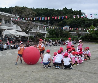 運動場で、赤白帽をかぶった大勢の子どもたちがトラック内に座り、その前のトラックを2人の子供が赤い大きな玉を転がして競技をしている運動会での写真