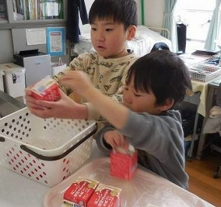 園児と思われる2人の男の子が、赤い小さな牛乳パックのような箱を、プラスチックの白いカゴやトレイに仕分けたり運んだりする手伝いをしている様子の写真