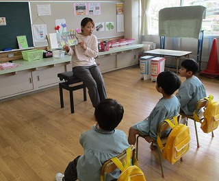 保育園の教室で、座って絵本を読み聞かせている女性教師に対し、水色のスモック姿の3人の園児たちが、椅子に座って真剣に耳を傾けている様子の写真