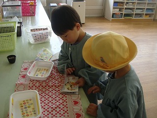 保育園の室内で、水色のスモックを着た2人の男の子が、チェック柄のテーブルクロスの上で、絵や文字のシールが貼られた容器や本を覗き込み、静かに活動に取り組んでいる写真