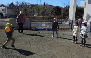 青空のもと、広場で、黄色の帽子をかぶった男の子が大きなオレンジ色のボールを両手で持って構え、ピンクの帽子をかぶった女の子たちと、それを見守る保育士らしき大人が、楽しそうにボール遊びをしている写真