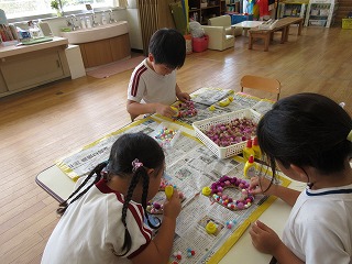 体操服姿の3人の園児たちが、床に置かれたテーブルの上で新聞紙を敷き、小さなポンポンや飾りをボンドで貼り付けて、輪っか状のリースのような制作活動に集中して取り組んでいる写真