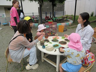 園庭と思われる屋外で、白丸いテーブルを囲み、教師と保護者、そしてピンクや黄色の帽子をかぶった子どもたちが、お皿に盛りつけた土や草などを使って楽しそうに泥遊びやおままごとをしている写真