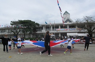 運動場で、先生や保護者と思われる大人と子どもたちが、紅白青のパラバルーンを広げて体操している様子を、校舎を背景に捉えた運動会の写真