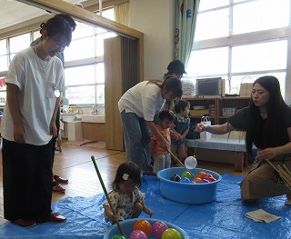 ビニールシートがひかれた室内の青いタライに入った水風船を、複数人の保育士らしき大人と、浴衣を着た女の子を含む園児たちが、釣り竿を使ってすくい上げるお祭り遊びを楽しんでいる写真