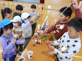 複数の園児たちが小学生の男の子2人がいる机を囲み、ストローや紙コップ、松ぼっくりなどの様々な材料を使って手作りおもちゃで遊んでいる様子の写真
