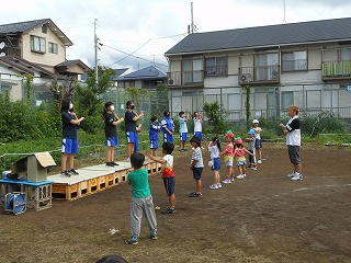 園庭で、いくつかのビールケースの上に板を載せた簡易的な台の上に立つ中学生達を見ながら、地面に並んで立つ複数の園児たちが体操をしている様子の写真