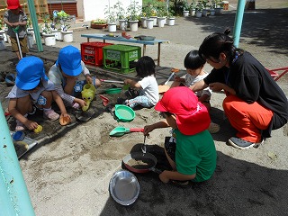 砂場で青い帽子や赤い帽子をかぶった園児たちや未就学児の子供達が先生に見守られながら、スコップやバケツなどのおもちゃを使って、砂を掘ったり盛ったり一緒に遊んでいる様子の写真