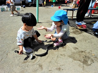 青い帽子をかぶった園児と未就学児の子供がしゃがみ込み、お玉とフライパンのようなおもちゃを使い、砂遊びをしている様子の写真