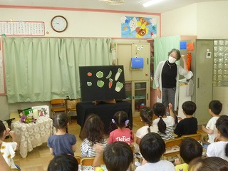先生が、黒い布を貼った台に様々な野菜のイラストを貼り説明しているのを園児たちが椅子に座り聞いている様子の写真