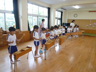 体操服を着た複数の園児たちが、2人の先生に見守られながら、等間隔に並べられた木琴や鉄琴などの楽器を演奏している様子の写真