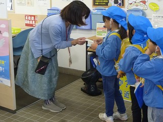 青い帽子をかぶった複数の園児たちが、郵便局の職員の女性に年賀状を手渡している様子の写真